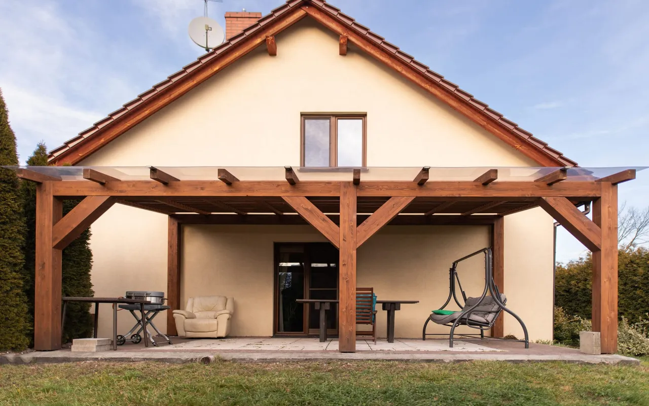 Drewniana pergola z przezroczystym dachem nad tarasem, obok grill i meble ogrodowe. Idealne miejsce na relaks.