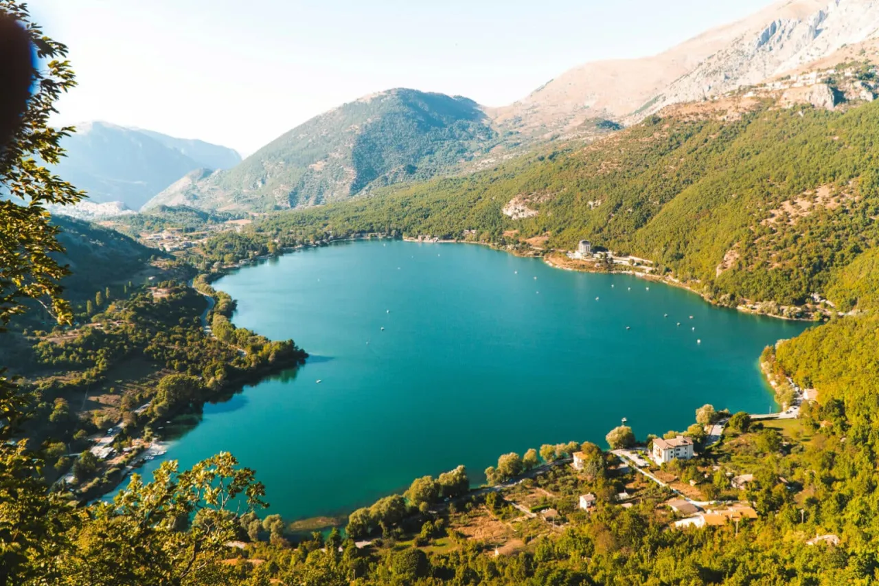 Malownicze jezioro otoczone zielonymi wzg&oacute;rzami, z widokiem na małe domki i g&oacute;ry. Idealne miejsce na wypoczynek w okolicach Pescary.