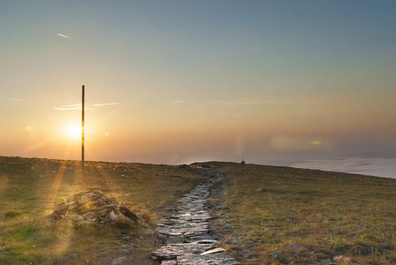 Kamienista ścieżka prowadzi przez trawiaste wzg&oacute;rza w kierunku zachodzącego słońca. W oddali widać mgłę, a na horyzoncie wieżę. To widok z hali barankowej bac&oacute;wki.