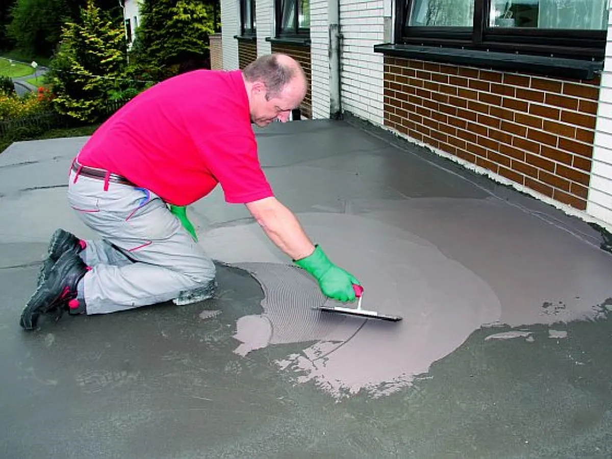 Mężczyzna w czerwonej koszulce i zielonych rękawicach nakłada zaprawę na taras.