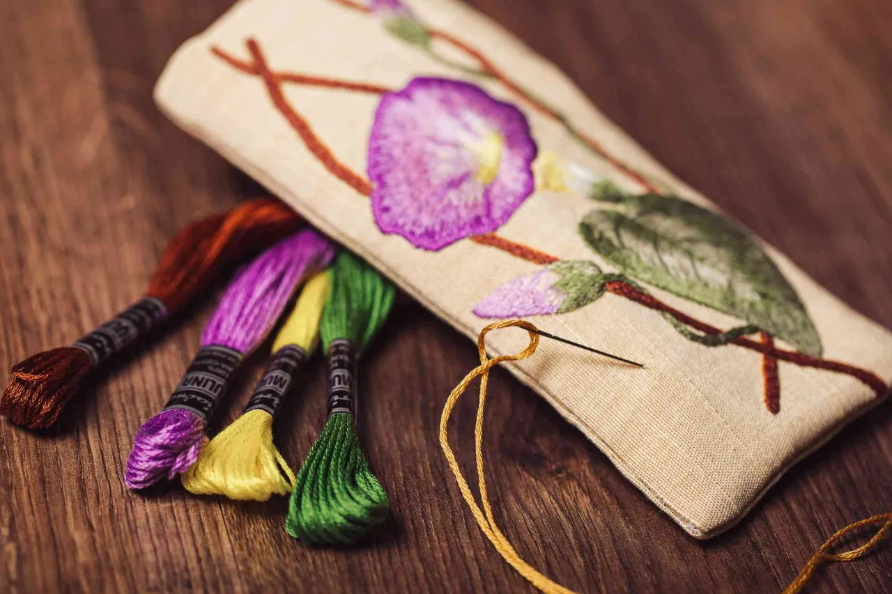 Kolorowe nici do haftowania obok haftowanego kwiatu na pł&oacute;tnie. Igła z ż&oacute;łtą nitką wchodzi w materiał.