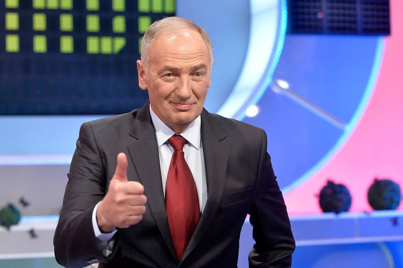 Uśmiechnięty mężczyzna w garniturze pokazuje kciuk w g&oacute;rę. W tle widać planszę z zielonymi polami, jak w teleturnieju Familiada.