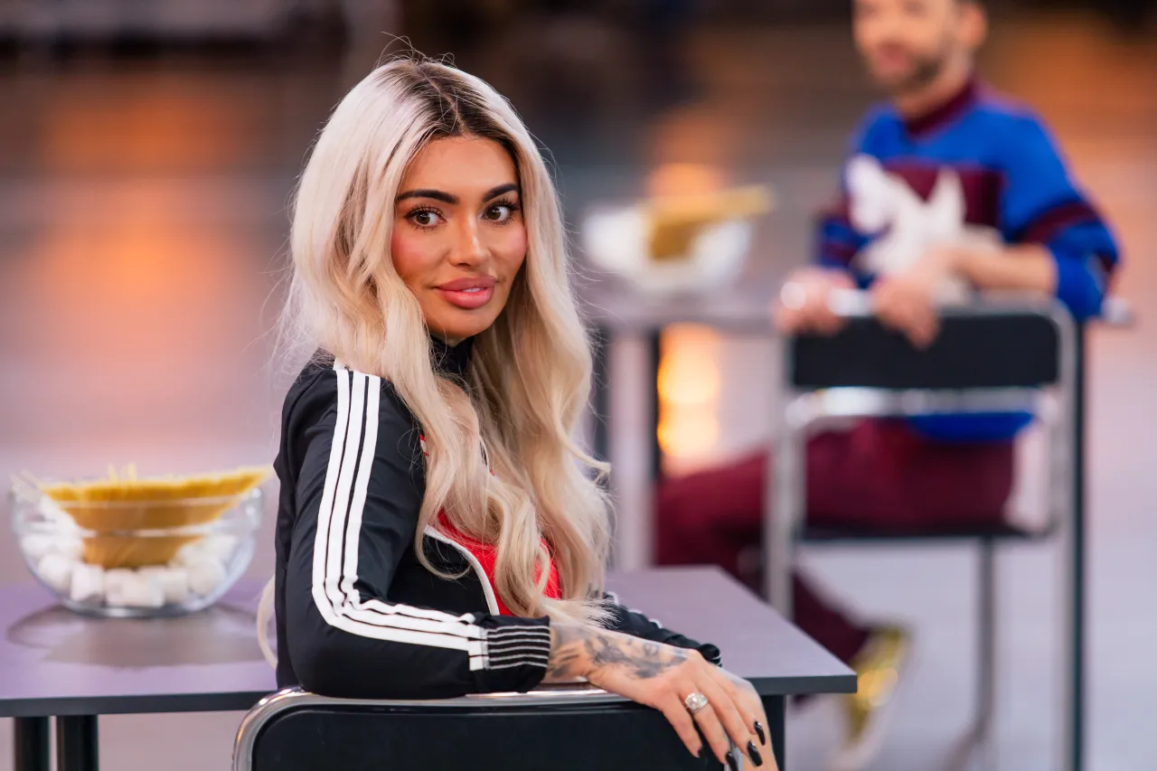 Ania z Królowych Życia w sportowym stroju z białymi pasami, z blond włosami i wyrazistym makijażem, siedzi przy stole z makaronem i cukrem.