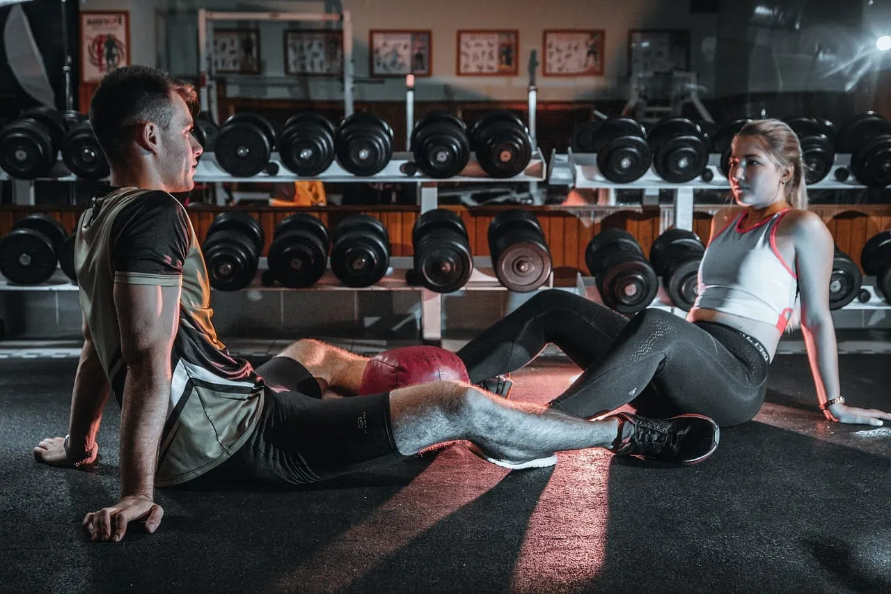 Para, siedząc na siłowni, dyskutuje o tym, ile cardio na redukcji jest optymalne. W tle stojaki z hantlami.