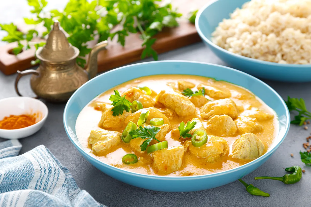 Aksamitny kurczak po tajsku w kremowym sosie, ozdobiony świeżą kolendrą i papryczką chili. Obok miseczka ryżu i przyprawy.