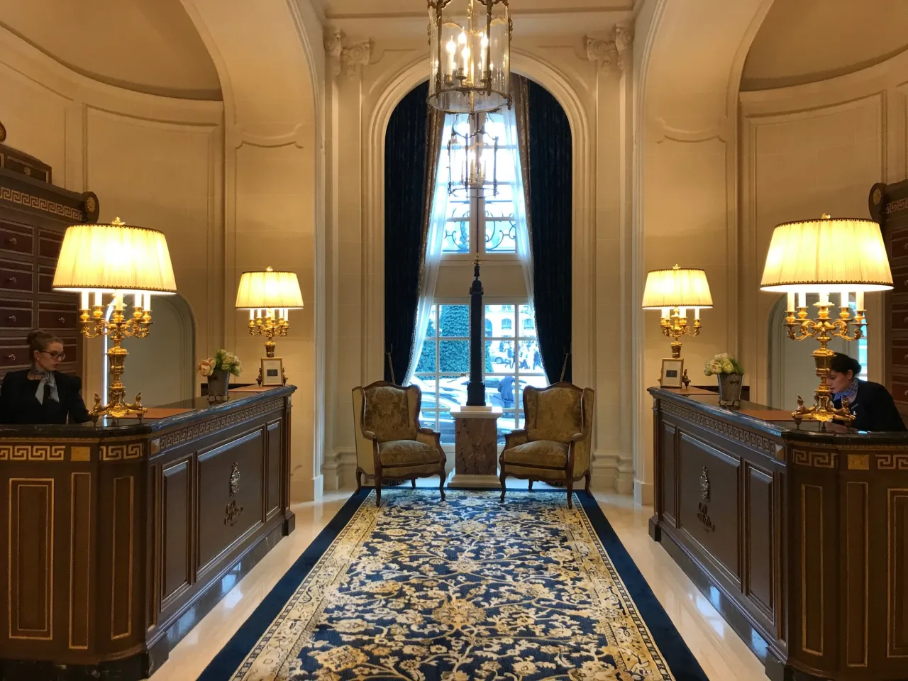 Eleganckie wnętrze hotelu Ritz w Paryżu z recepcjonistkami przy luksusowych ladach, ozdobnym dywanem i stylowymi fotelami.