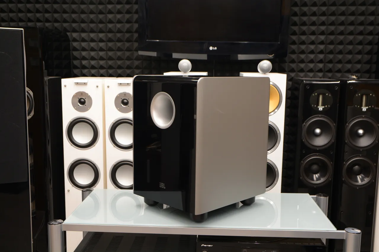 Czarny, błyszczący jbl subwoofer aktywny estradowy z szarym panelem, otoczony innymi głośnikami.