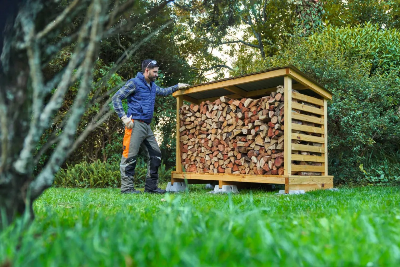Mężczyzna w niebieskiej kamizelce stoi przy drewutni pełnej drewna opałowego. Czas schnięcia drewna opałowego zależy od wielu czynników.