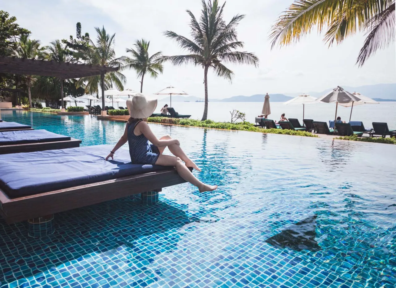 Kobieta w kapeluszu odpoczywa przy basenie z widokiem na morze. Idealne miejsce, gdzie rezerwować noclegi na wakacje marzeń.