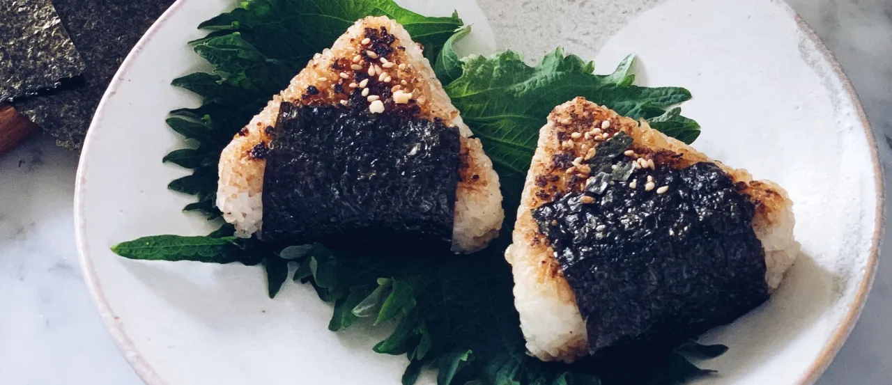 Dwa pyszne yaki onigiri, ryżowe tr&oacute;jkąty z nori i sezamem, podane na liściach shiso.