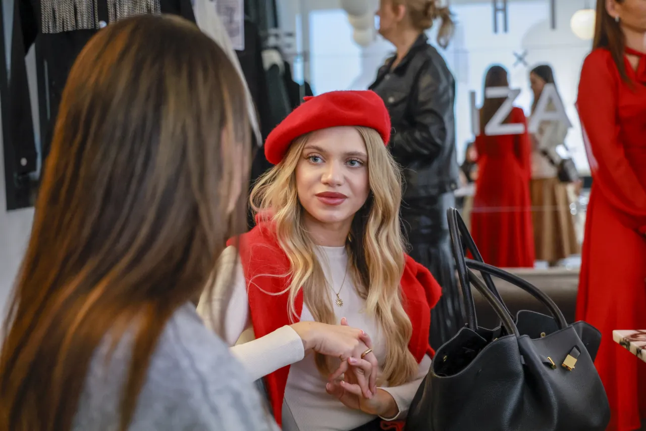 Dziewczyna w czerwonym beretku i sweterku zastanawia się, kto ubiera ją dzisiaj. W tle widać inne kobiety w modnych strojach.