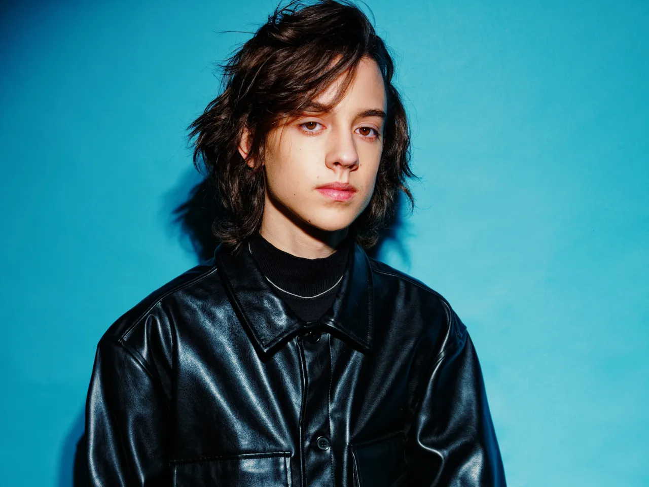 Młody mężczyzna w czarnej skórzanej kurtce, z burzą brązowych włosów, patrzy wprost, jakby czekał na znak, kto śpiewa.