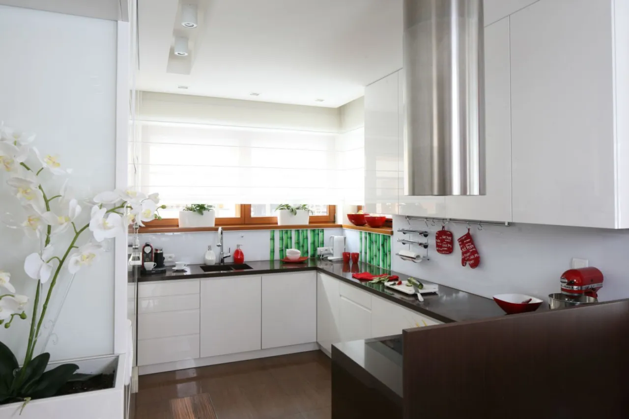 Nowoczesna kuchnia z białymi szafkami i czarnym blatem. Okna w kuchni znajdują się na standardowej wysokości, nad nimi białe rolety.