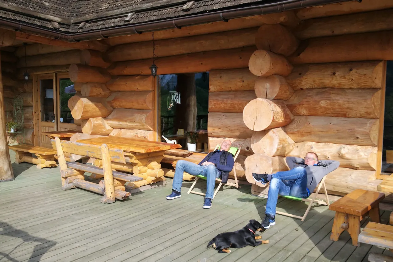 Dwóch mężczyzn odpoczywa na werandzie domu z bali, obok leży pies. Tyle drewna na dom z bali to marzenie wielu.