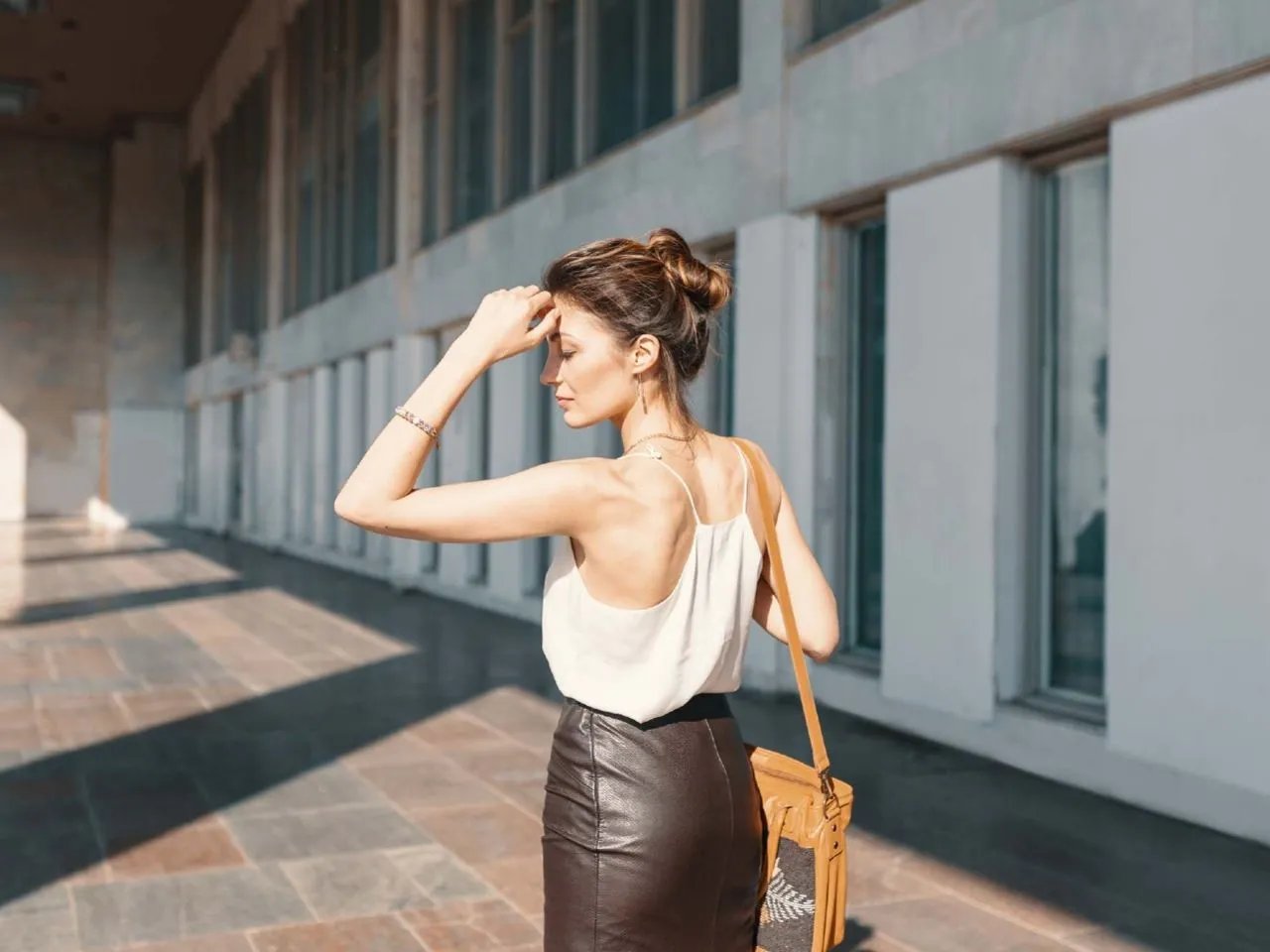 Elegancka kobieta w białej bluzce na ramiączkach i brązowej sk&oacute;rzanej sp&oacute;dnicy, zastanawia się, jaka bluzka do sk&oacute;rzanej sp&oacute;dnicy pasuje najlepiej.