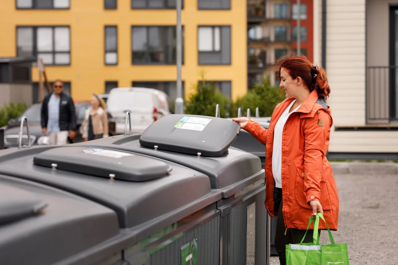 Kobieta z pomarańczową kurtką otwiera pojemnik na odpady komunalne. W tle widać budynki i inne osoby.