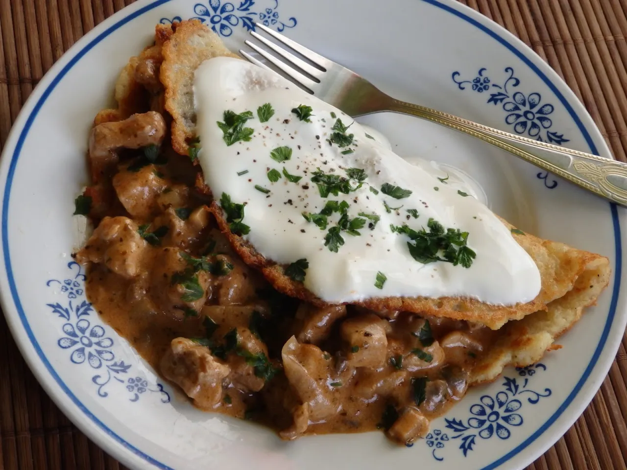 Placki po zbójnicku: chrupiące placki ziemniaczane z gulaszem mięsnym w sosie i kleksem śmietany, posypane natką pietruszki.