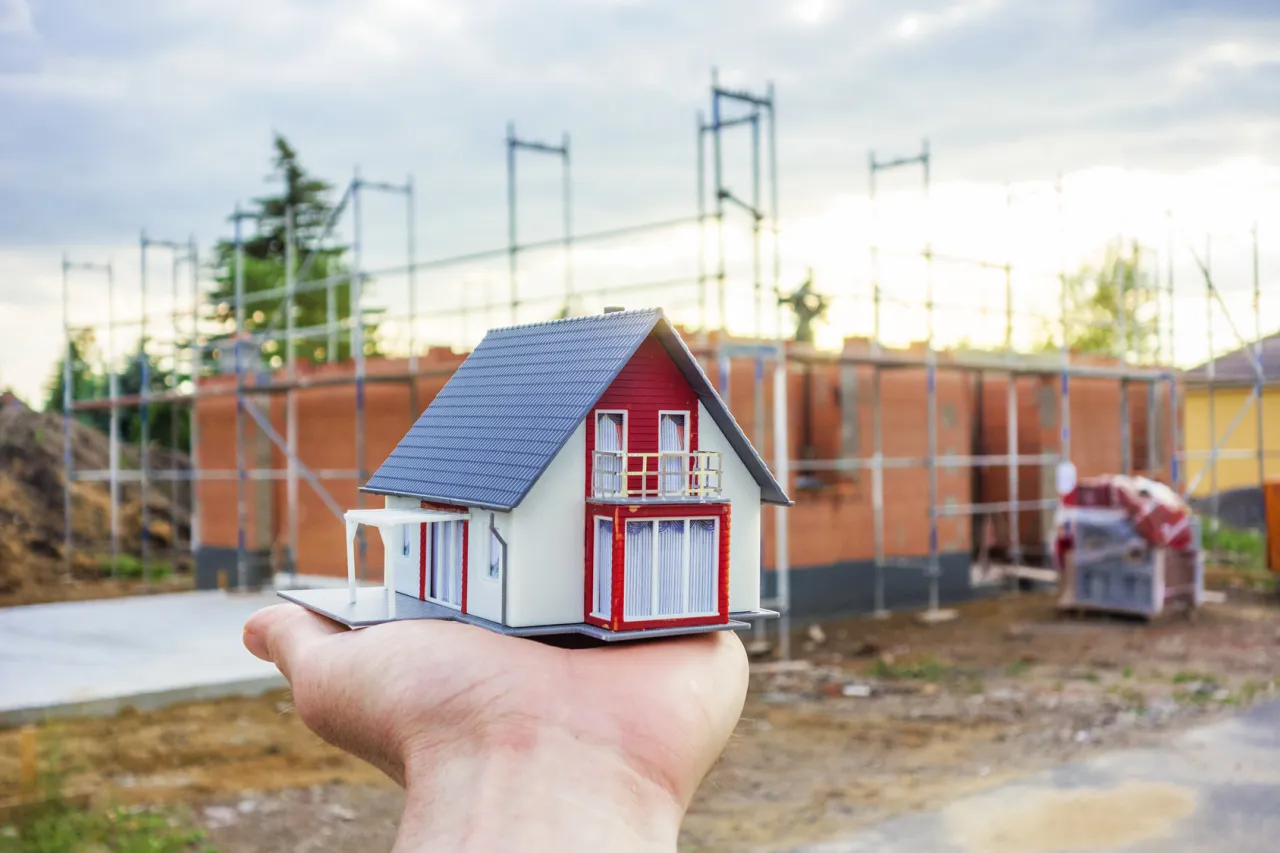 Model domu w ręce, w tle budowa. Pomyśl o ubezpieczeniu domu w budowie, by chronić swoje marzenia.