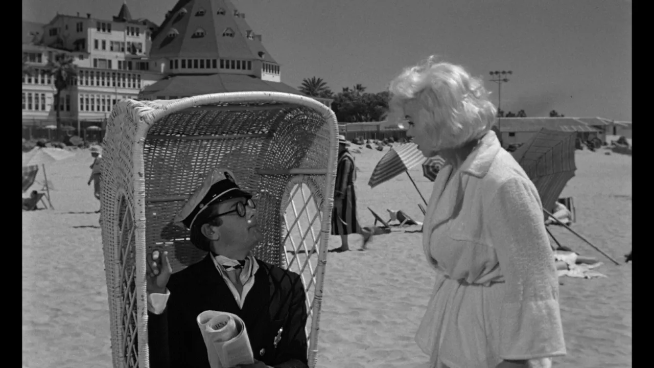 Pół żartem pół serio obsada: mężczyzna w kapeluszu kapitana siedzi w wiklinowym fotelu, rozmawiając z kobietą w szlafroku na plaży.