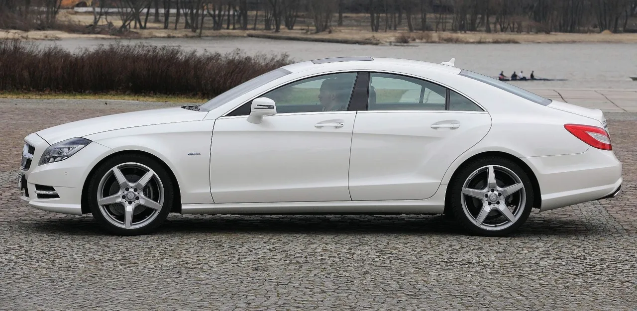 Biały Mercedes-Benz CLS 63 AMG z przyciemnianymi szybami, zaparkowany na brukowanej drodze, z widokiem na jezioro i drzewa w tle.