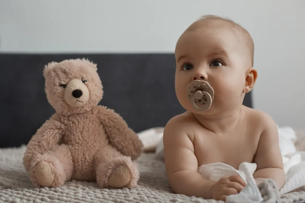 Urocze niemowlę z uspokajającym smoczkiem, obok pluszowy miś. Dziecko wygląda na zrelaksowane, choć może lekko się poci.