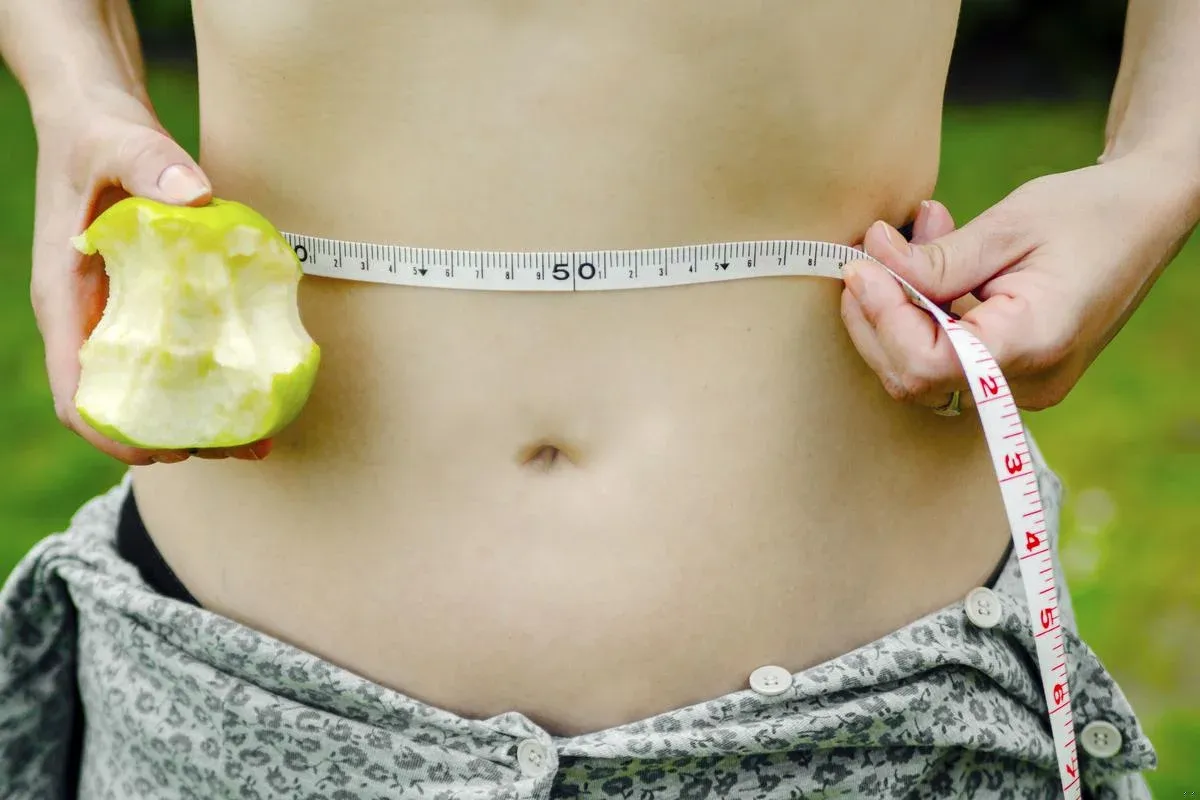 Kobieta mierzy talię centymetrem, trzymając nadgryzione jabłko. To dobry początek, by dowiedzieć się, jak zacząć redukcję.