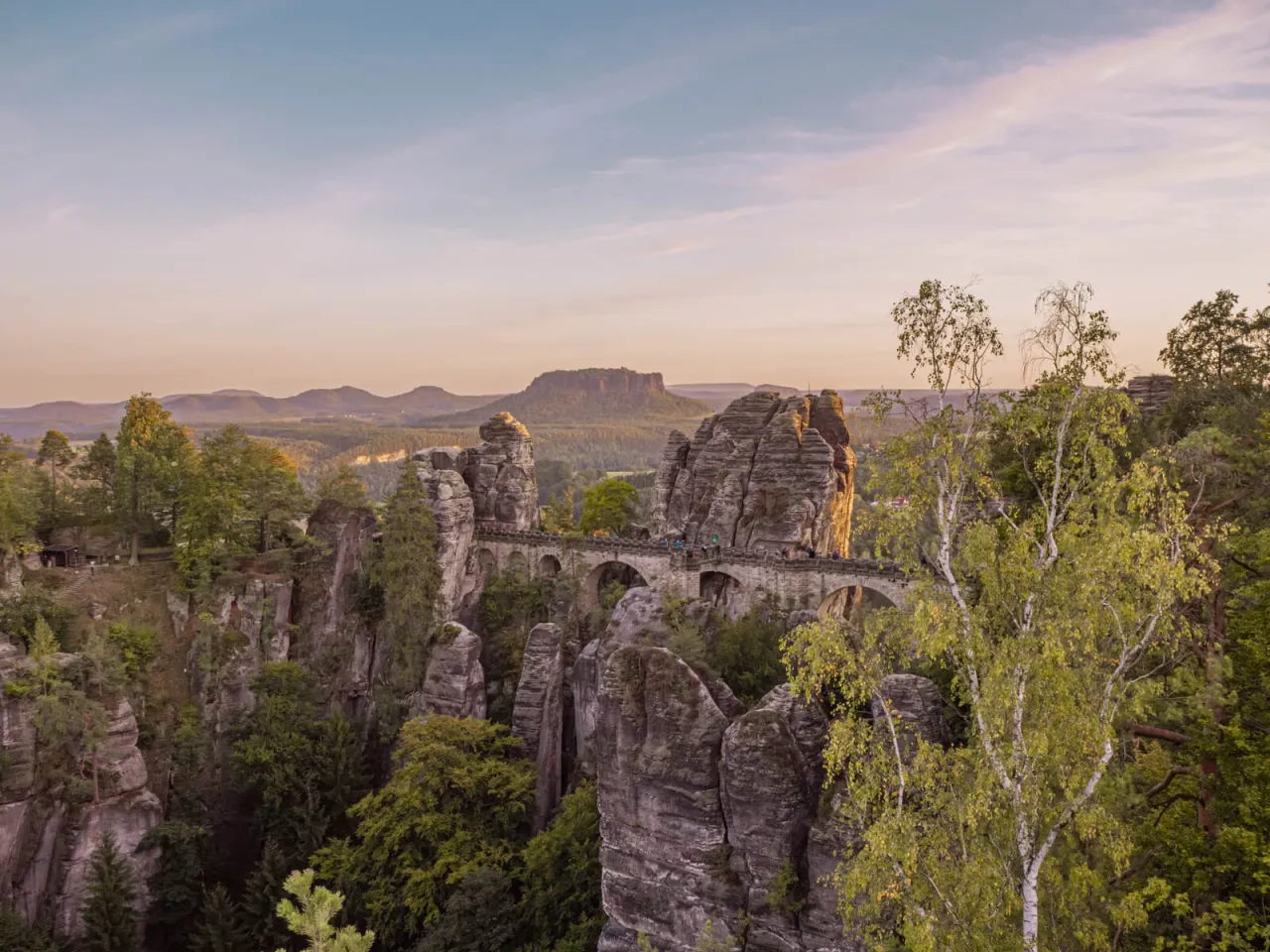 Kamienny most Bastei w Szwajcarskiej Saksonii, otoczony skalnymi formacjami i zielenią, z widokiem na g&oacute;ry w oddali.