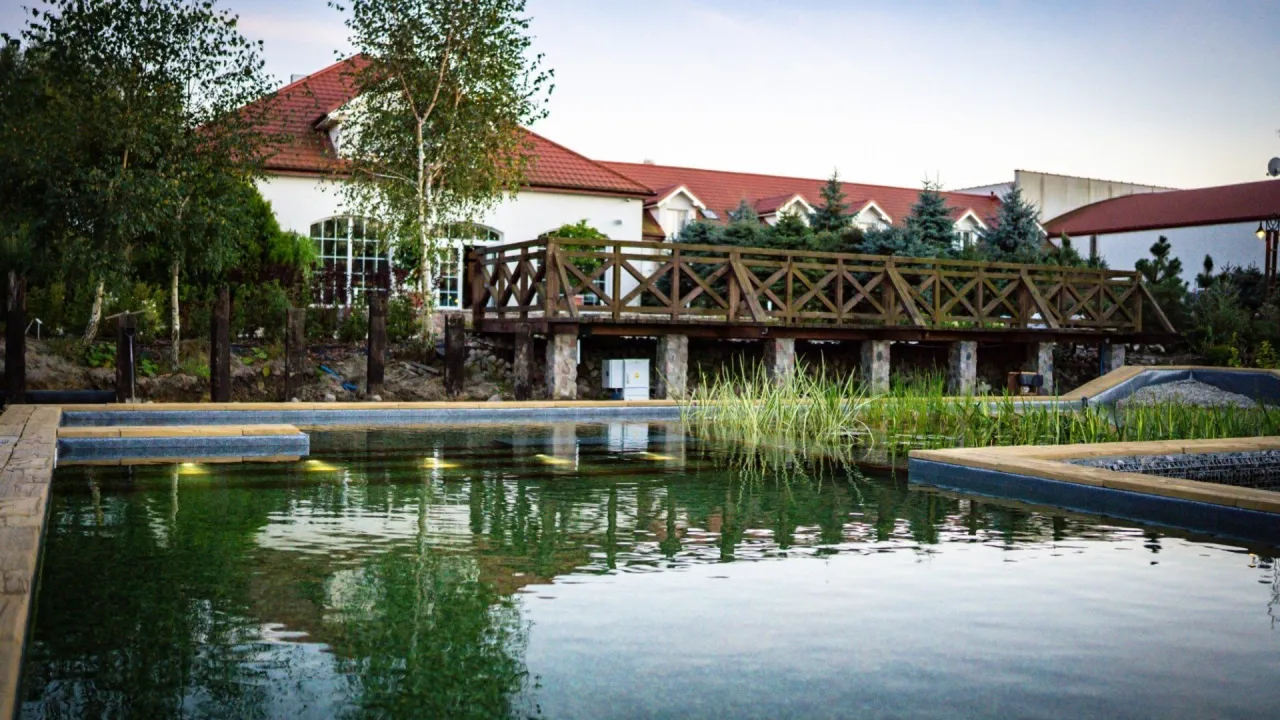 Relaks w spa Białystok i okolice: malowniczy staw, drewniany mostek i budynek z czerwoną dachówką.