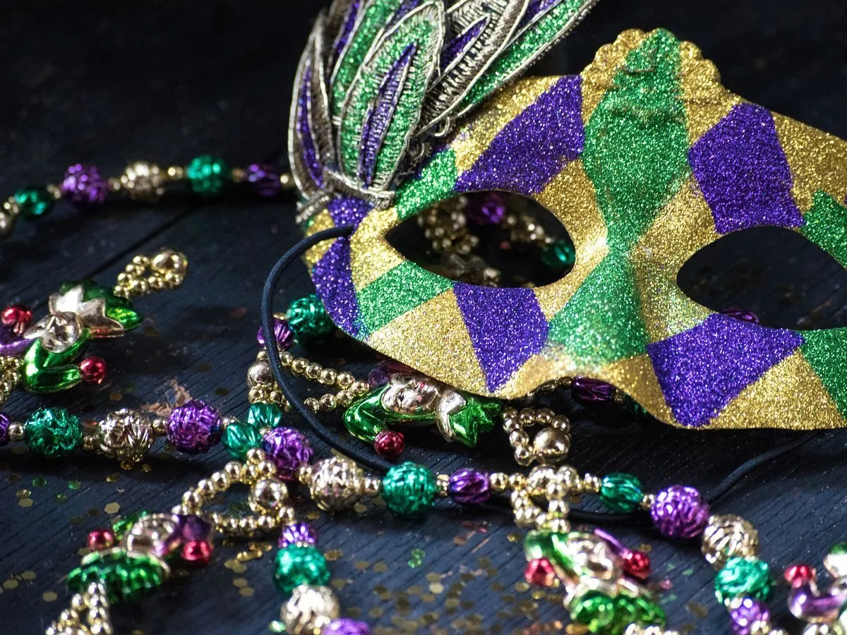 Błyszcząca maska karnawałowa w kolorach fioletu, zieleni i złota, ozdobiona pi&oacute;rami, leży obok naszyjnik&oacute;w z koralik&oacute;w. Idealna inspiracja, jak zrobić maskę karnawałową.