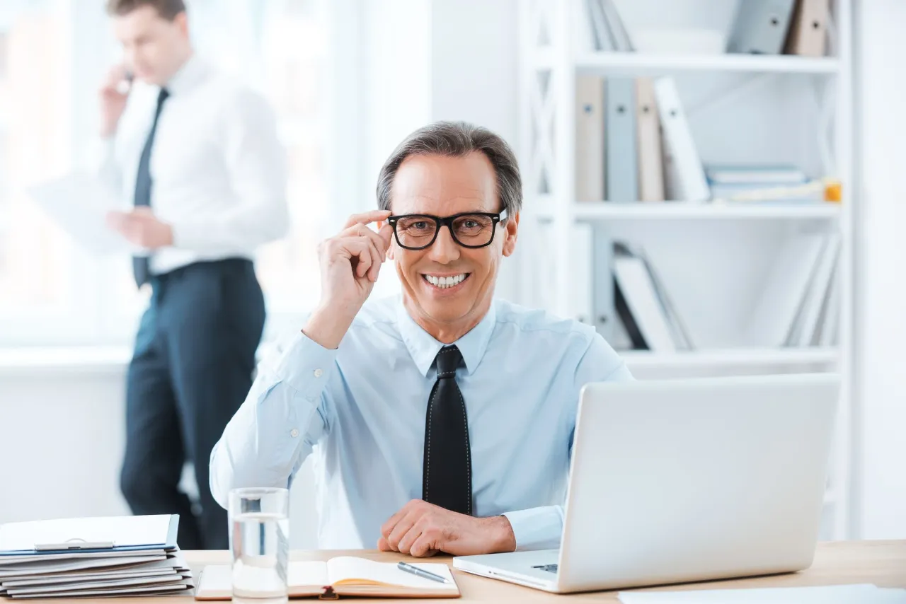 Uśmiechnięty biznesmen poprawia okulary, gotowy do rozliczenia zwrotu za okulary na kogo faktura. Obok laptop i dokumenty.