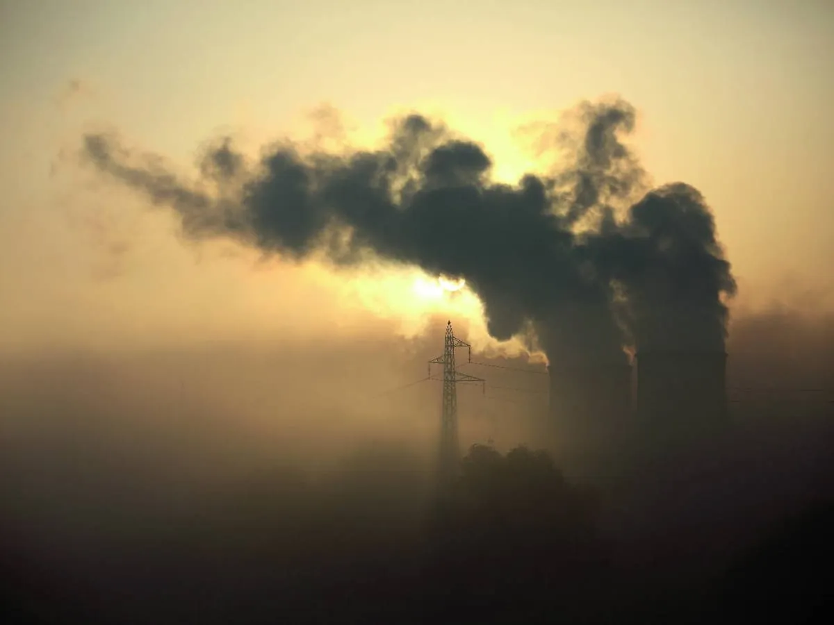 Dymiące kominy elektrowni w mglisty poranek. Czy smog ma zapach? Słońce przebija się przez kłęby dymu, a w oddali widać linię energetyczną.