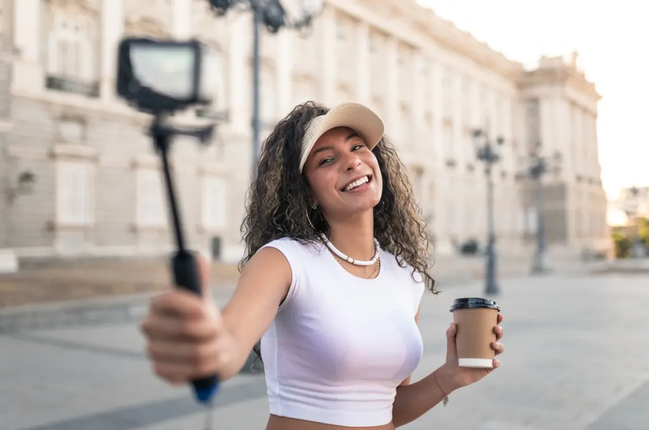 Uśmiechnięta content creator z kawą w ręku nagrywa vlog na selfie sticku przed piękną budowlą.