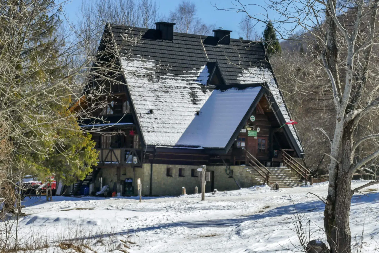 Zimowa sceneria wok&oacute;ł bac&oacute;wki pod Małą Rawką. Drewniany budynek z ciemnym dachem pokrytym śniegiem, otoczony nagimi drzewami.
