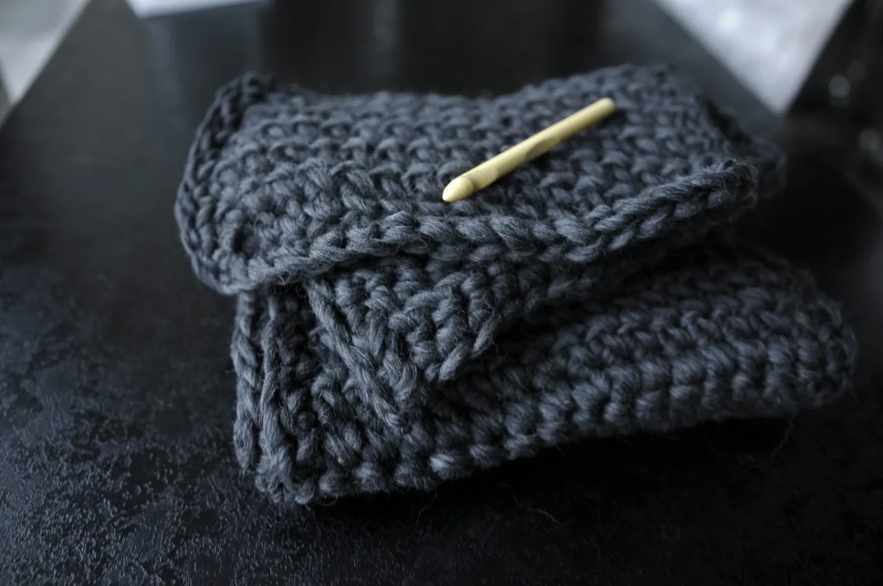 Gruby, ciemnoszary szalik z wełny czesankowej, na kt&oacute;rym leży drewniana szydełko.