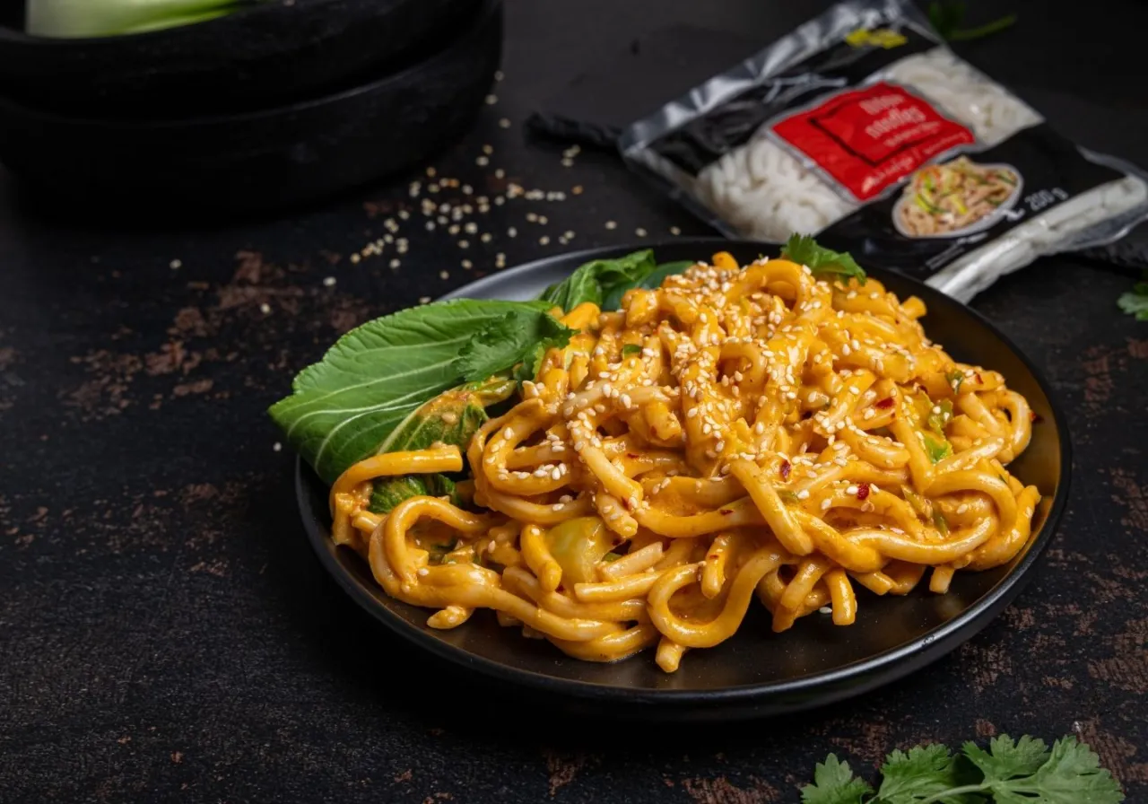 Grube, pomarańczowe kluski biang biang w sosie, posypane sezamem i chili, z zielonym pak choi na talerzu.