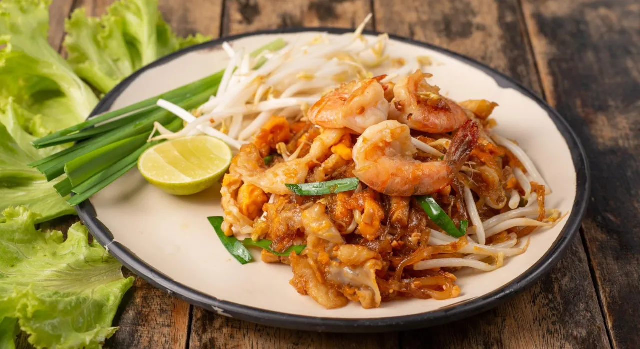 Pyszne tajskie jedzenie: Pad Thai z krewetkami, kiełkami fasoli mung, szczypiorkiem i ćwiartką limonki na talerzu.