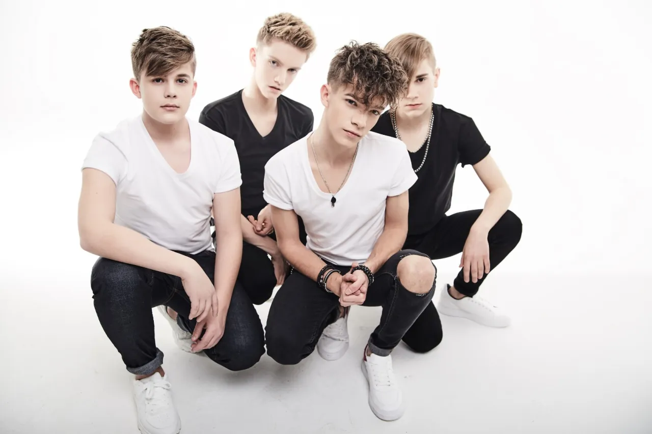 Czterech młodych mężczyzn w białych i czarnych koszulkach kuca na białym tle. Zastanawiasz się, ile trwa koncert 4dreamers?