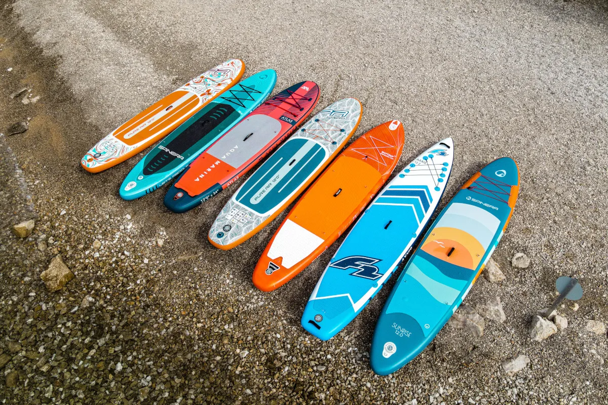 Siedem dmuchanych desek do pływania w r&oacute;żnych kolorach i wzorach leży na kamienistej plaży, gotowych na wodne przygody.