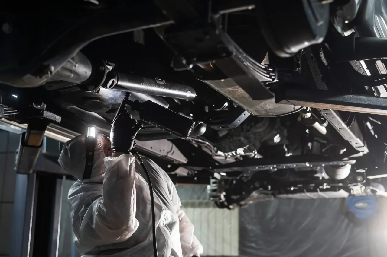 Pracownik w kombinezonie i masce wykonuje konserwację podwozia bez kanału, używając pistoletu do natrysku.