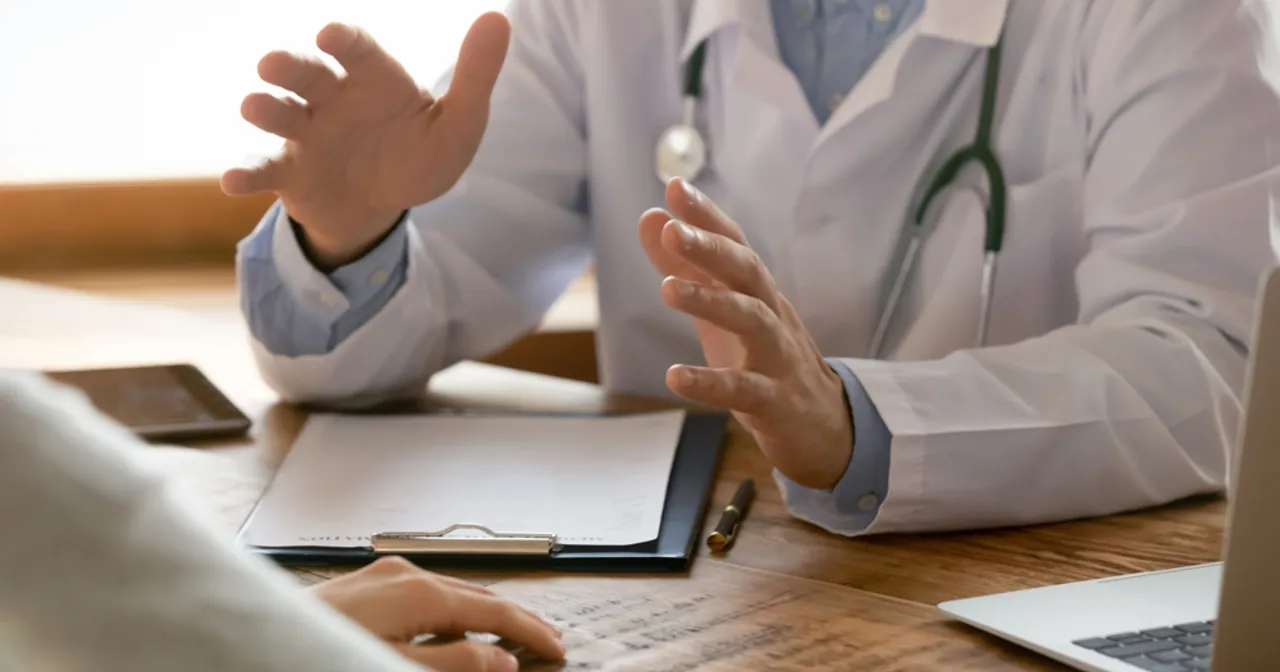 Lekarz w kitlu tłumaczy coś pacjentce, gestykulując. Na stole dokumenty, laptop i telefon. Taki lekarz nie może poślubić swojej pacjentki.