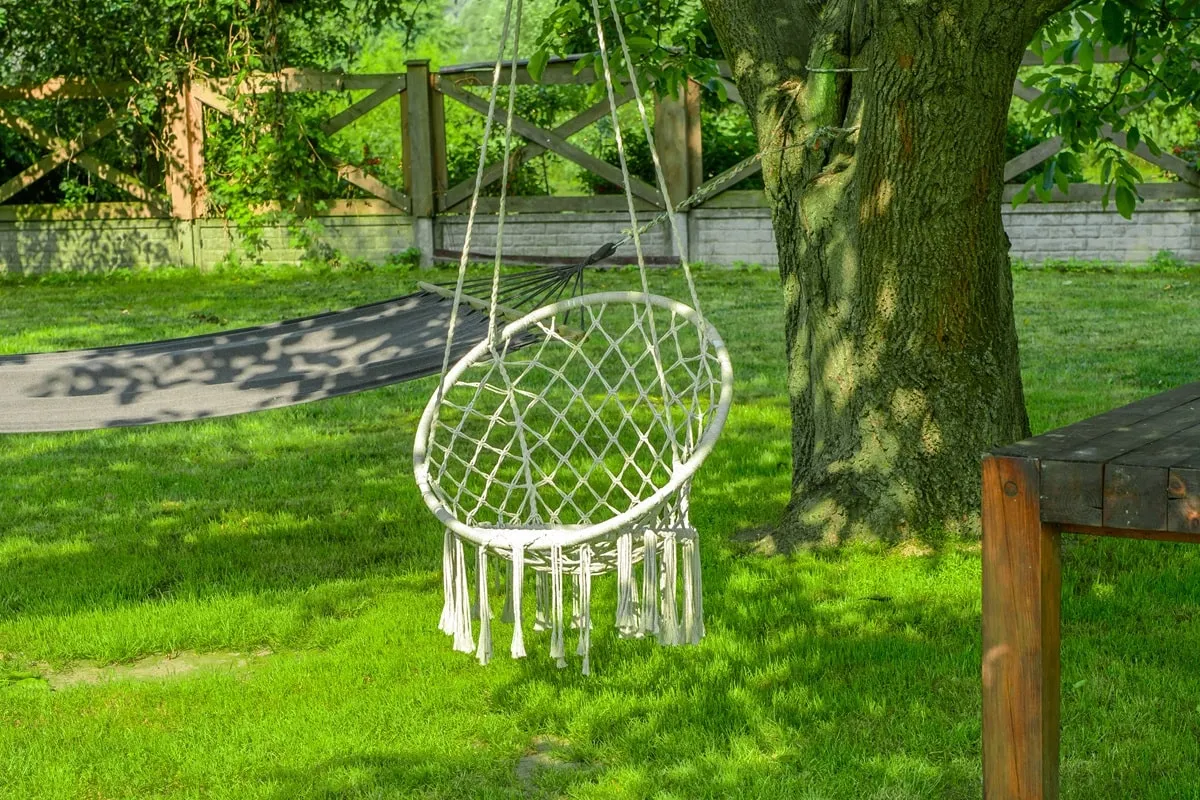 Oto jak zrobić huśtawkę ogrodową: biała, pleciona huśtawka z frędzlami wisi na drzewie, obok hamaka i stołu.