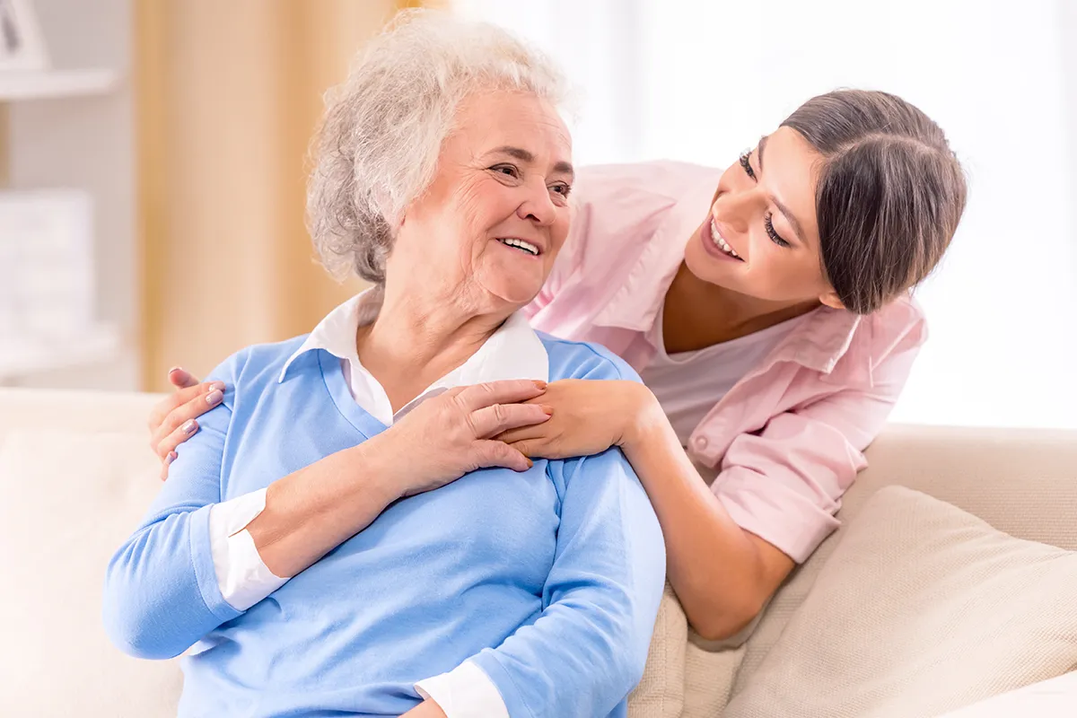 Uśmiechnięta młoda kobieta obejmuje starszą panią, która szuka rozwiązań co na nietrzymanie moczu u starszej osoby.