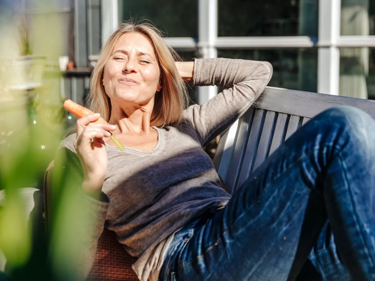 Kobieta po 50-tce z marchewką w ręku, relaksująca się na ławce. Szuka odpowiedzi na forum: jak schudnąć po 50?