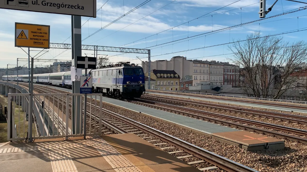 Pociąg TLK zbliża się do stacji. Na peronie widać znaki ostrzegawcze, a w tle budynki mieszkalne.