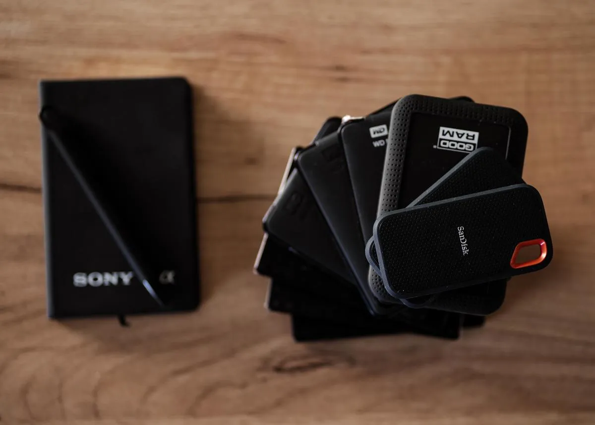Na drewnianym blacie leży czarny notatnik Sony i stos zewnętrznych dysk&oacute;w twardych, na kt&oacute;rych fotograf przechowuje zdjęcia.