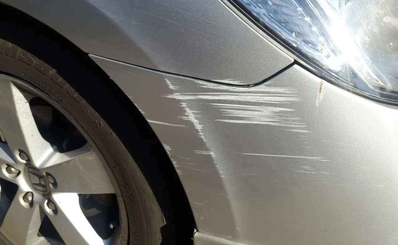 Srebrny błotnik samochodu z licznymi zarysowaniami, śladami po kolizji. Sprawca szkody z OC pewnie będzie musiał pokryć koszty naprawy.