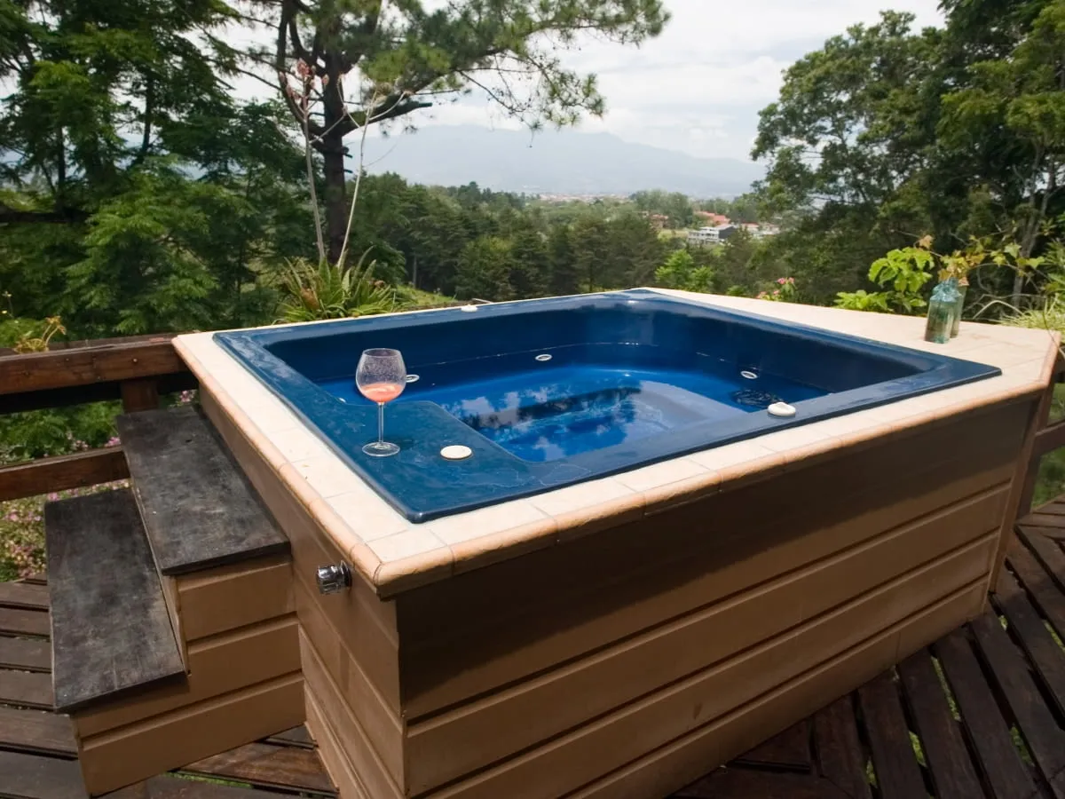 Jak zabudować jacuzzi ogrodowe? Widok na taras z niebieskim jacuzzi, schodkami i kieliszkiem różowego napoju. W tle malowniczy krajobraz z górami.