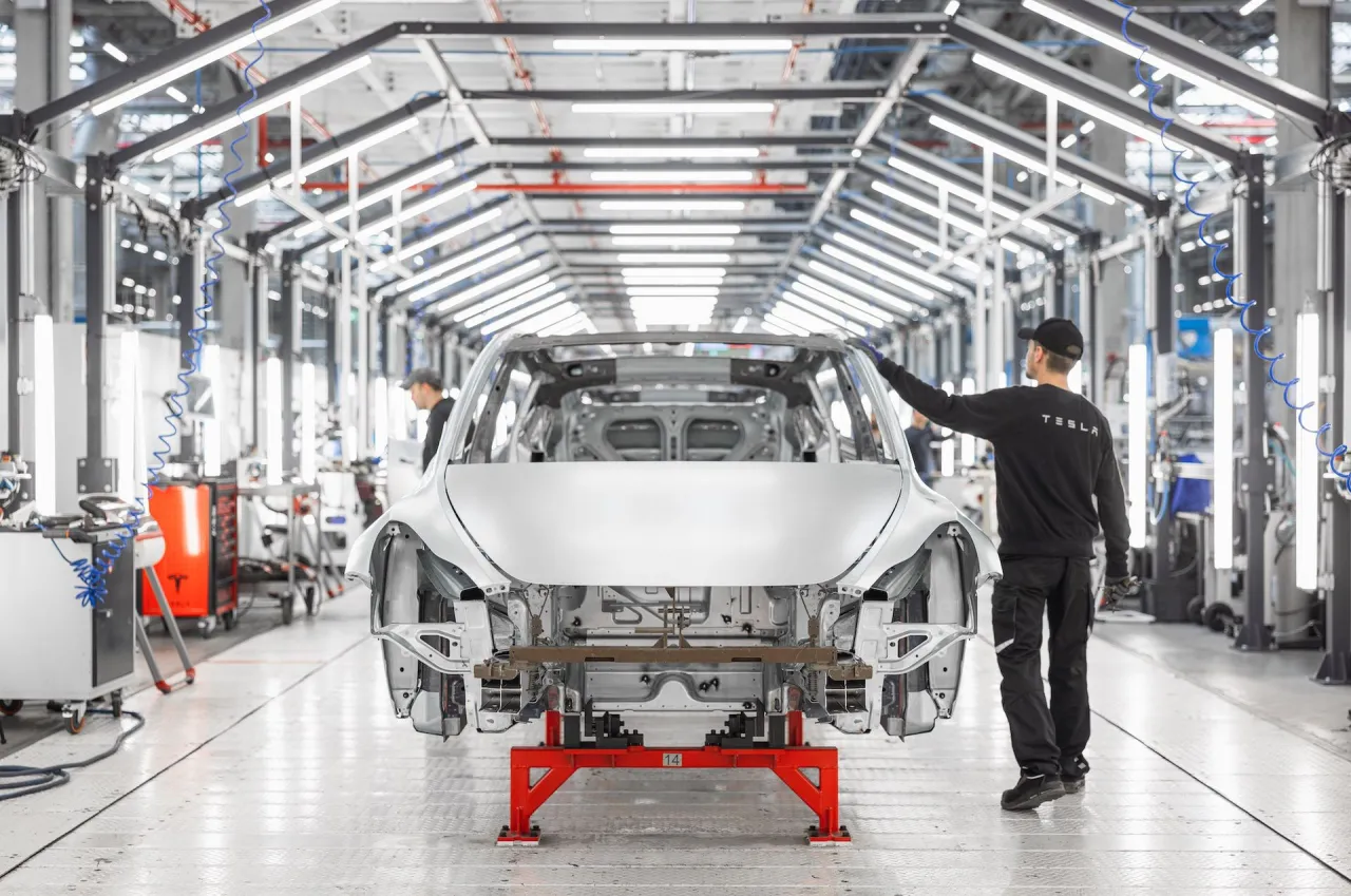 Pracownik w fabryce Tesli pracuje nad nadwoziem samochodu. Tesla kto produkuje? Odpowiedź jest widoczna na jego bluzie.