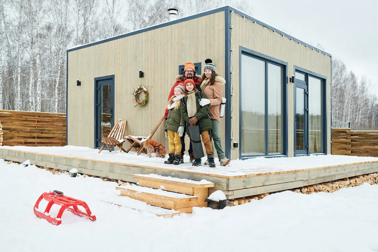 Rodzina pozuje przed swoim całorocznym domkiem letniskowym w zimowej scenerii. Na śniegu leży czerwone sanki.