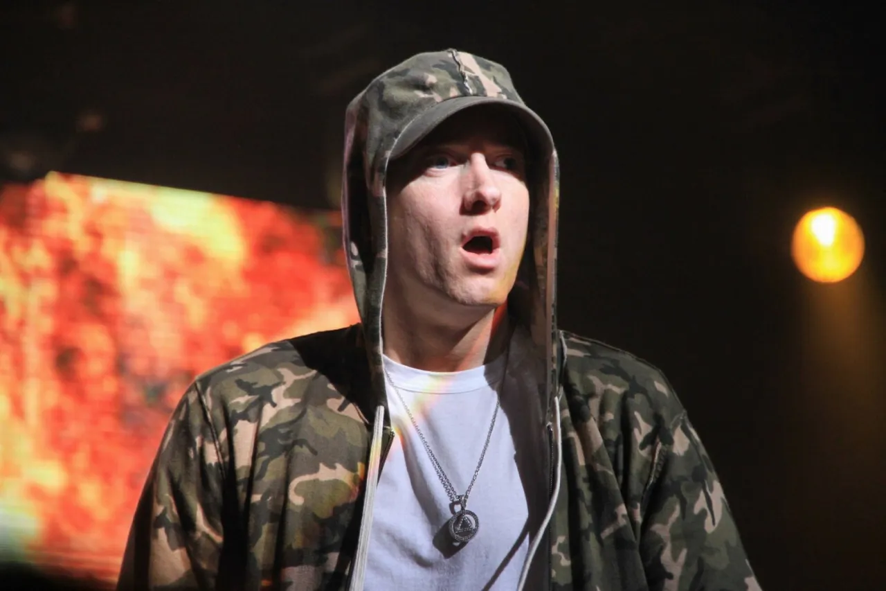 Eminem w bluzie z kapturem i naszyjnikiem, z otwartymi ustami. W tle płonący ekran. Ciekawe, ile ma lat Eminem?